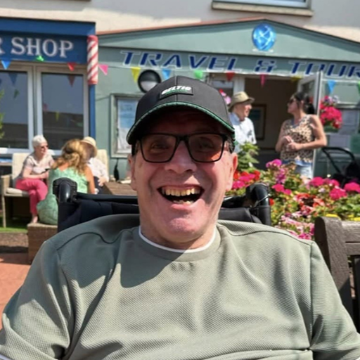 Care home resident David has a green sweat shirt and a dark cap on. He is smiling. Behind him are some residents and families of the care home, with the gardens shop front design visible in the distance. 
