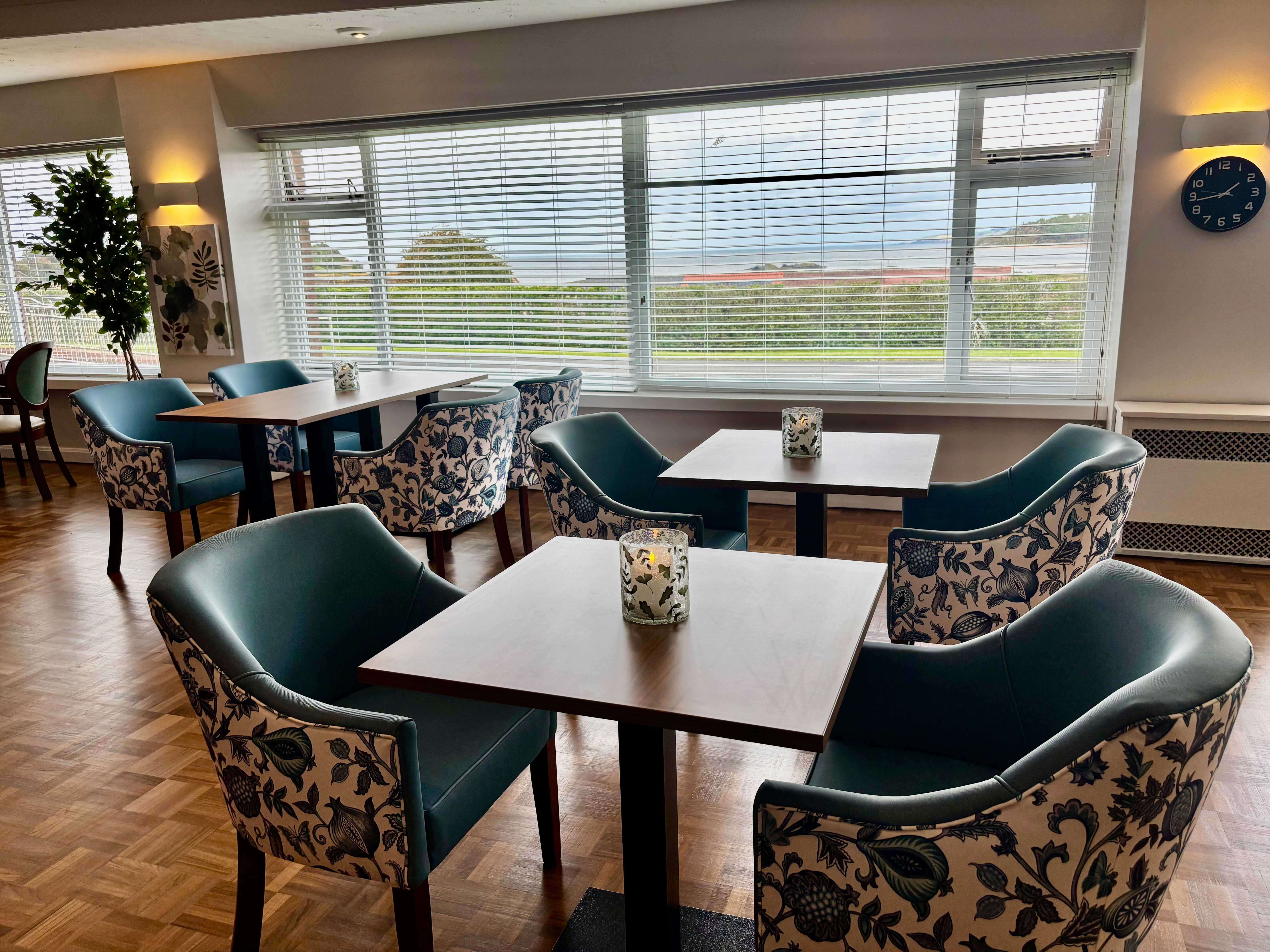 A bright, modern bistro seating area with several small dark wood tables and comfortable chairs upholstered in solid teal blue and a coordinating blue-and-white floral pattern. Large windows overlook a grassy area and the sea.