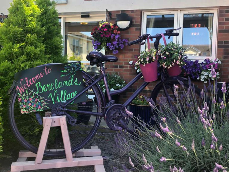 Berelands Village: A bike sits in amongst a garden area with a sign reading ' Welcome to Berelands Village ' 