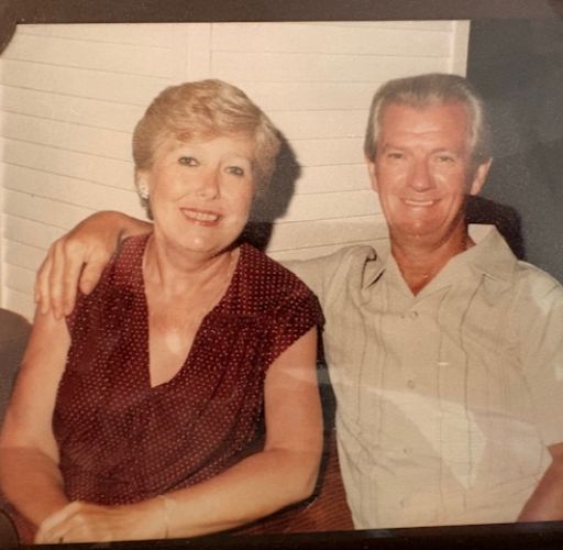 A woman and a man sit together. On the left, the woman has short blonde hair and is wearing a dark red polka-dot blouse. The man, on the right, has greyish-white hair and is wearing a light-coloured, short-sleeved button-down shirt with his arm around the woman's shoulder. Both are smiling warmly at the camera.