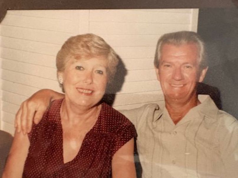 A woman and a man sit together. On the left, the woman has short blonde hair and is wearing a dark red polka-dot blouse. The man, on the right, has greyish-white hair and is wearing a light-coloured, short-sleeved button-down shirt with his arm around the woman's shoulder. Both are smiling warmly at the camera.