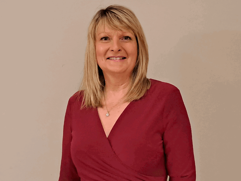 Yvonne Richardson has blonde hair and is wearing a red dress. She is standing infront of a white backdrop.
