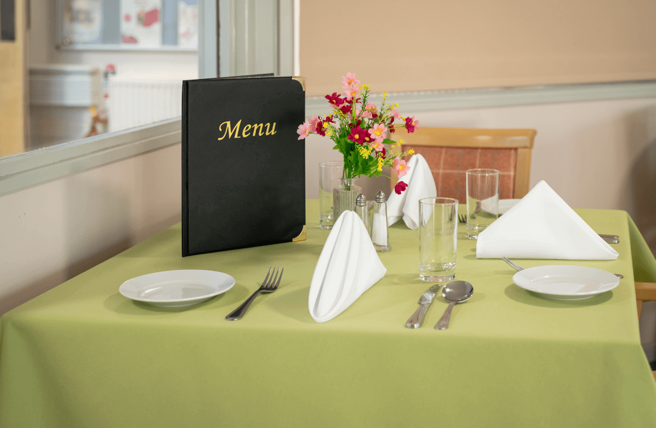 In the dining room, a table is prepared with cutlery, napkins, a floral centerpiece, and menus.