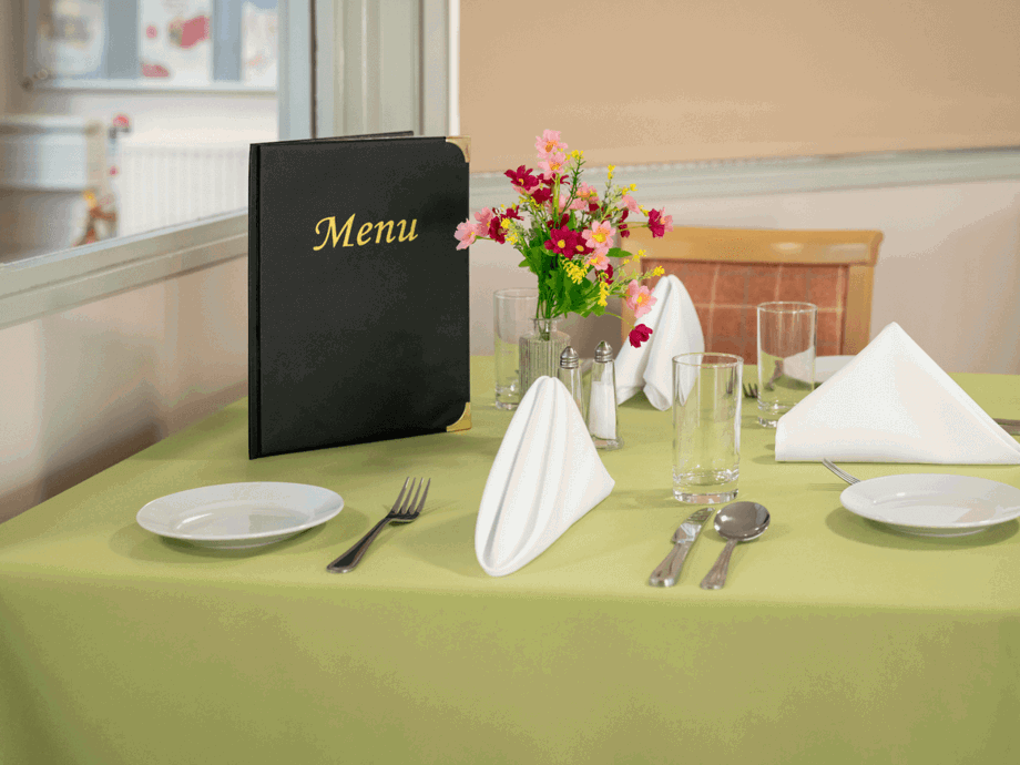 In the dining room, a table is prepared with cutlery, napkins, a floral centerpiece, and menus.