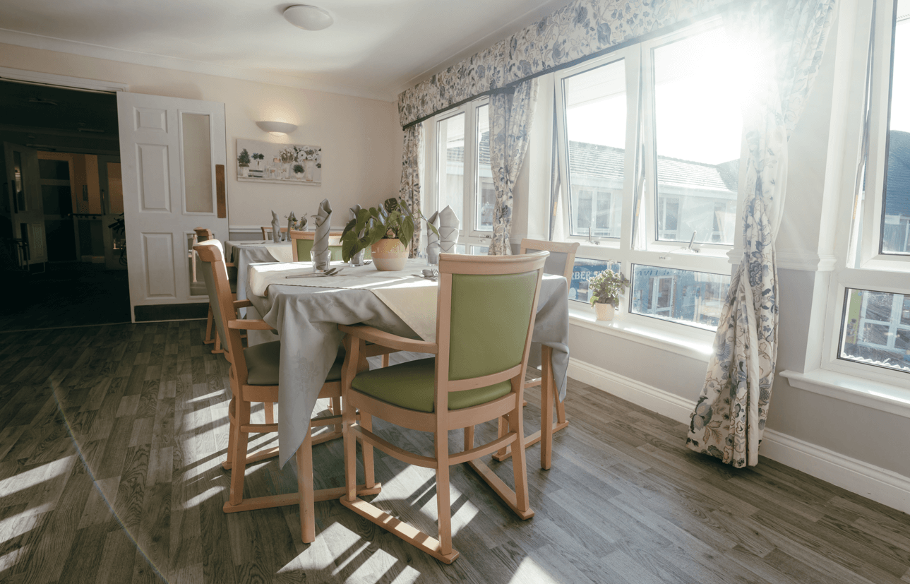 A bright, sunlit dining room with several tables set with grey tablecloths and green-backed chairs. Large windows with floral curtains line one wall, letting in significant natural light. A potted plant sits in the center of the foreground table.