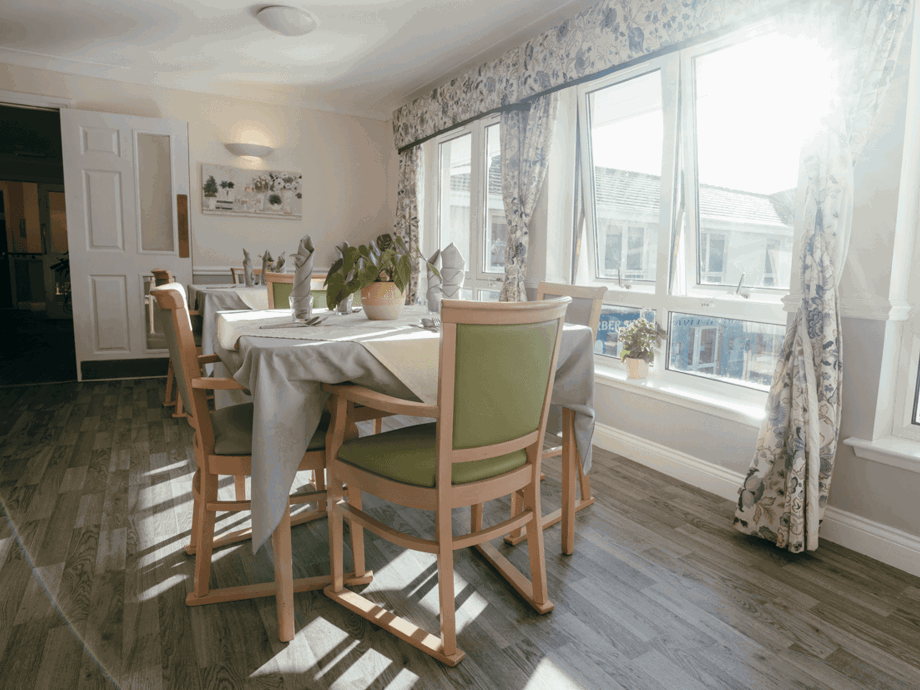 A bright, sunlit dining room with several tables set with grey tablecloths and green-backed chairs. Large windows with floral curtains line one wall, letting in significant natural light. A potted plant sits in the center of the foreground table.