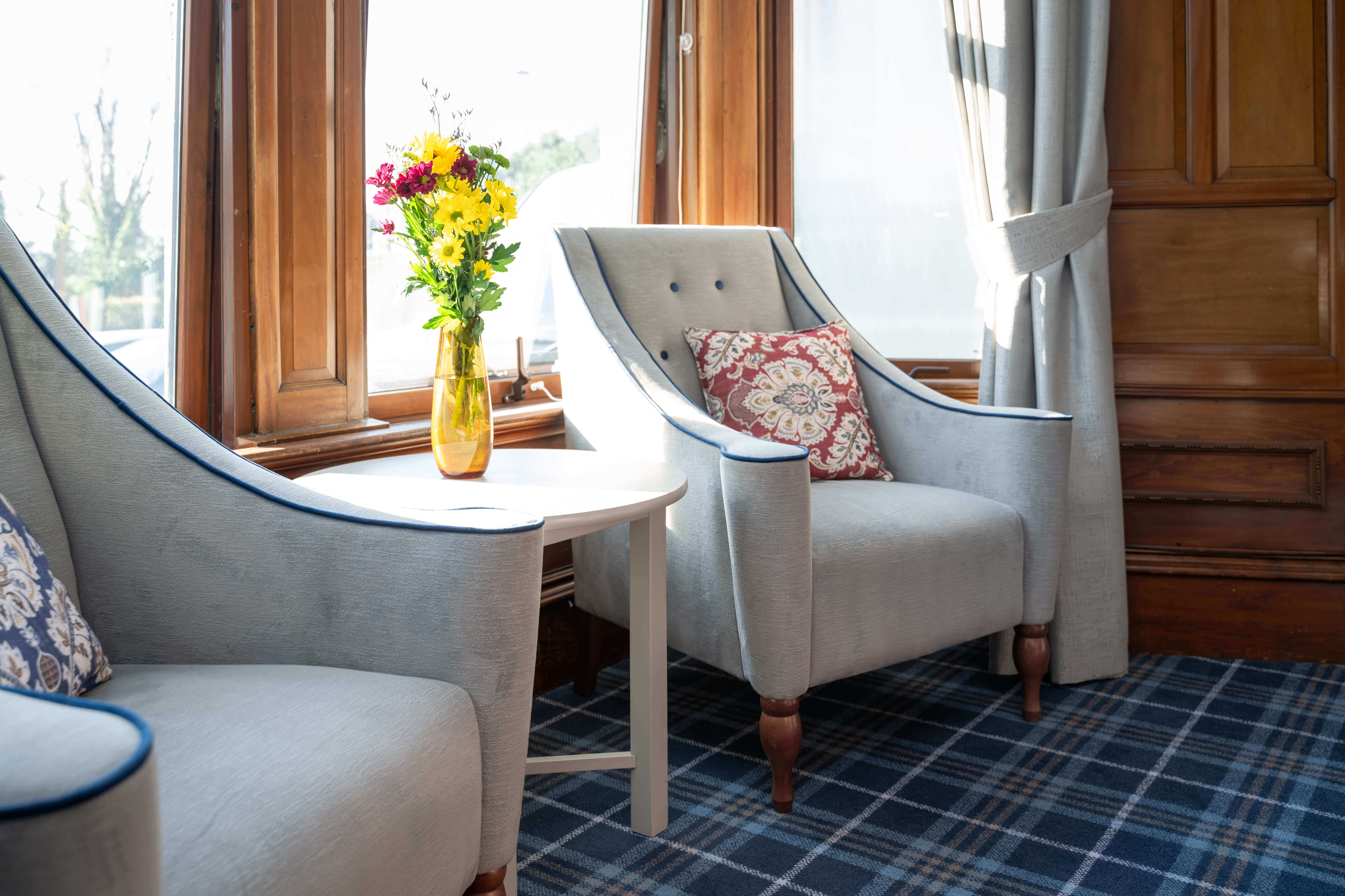 In the communal lounge, two armchairs and a table with flowers are positioned in front of a large window area.