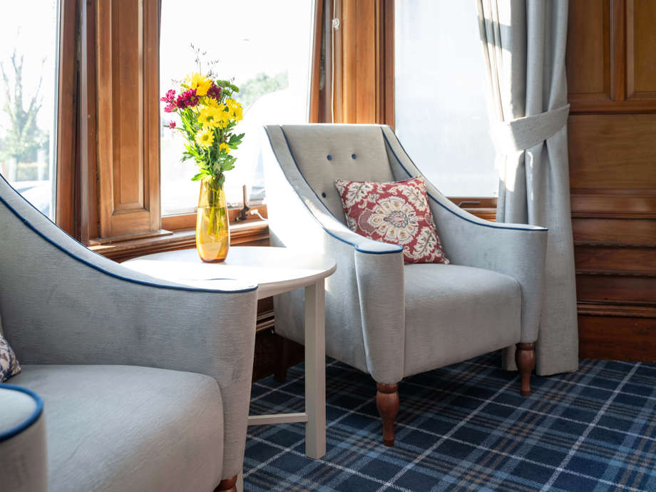 In the communal lounge, two armchairs and a table with flowers are positioned in front of a large window area.