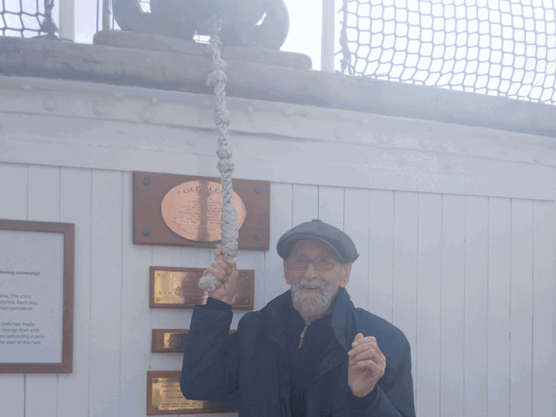 Care home resident Kenneth, smiling, rings a ship's brass bell mounted on a wooden stand
