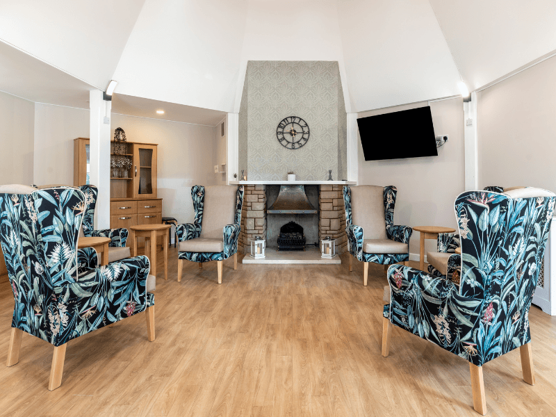 A spacious seating area centered around a stone fireplace with a decorative mantel and a large wall clock. Four high-backed wingback chairs with a dark tropical leaf pattern are arranged in a semi-circle on light wood flooring. A wall-mounted TV is positioned to the right of the fireplace.