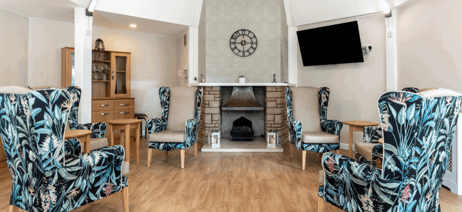 A spacious seating area centered around a stone fireplace with a decorative mantel and a large wall clock. Four high-backed wingback chairs with a dark tropical leaf pattern are arranged in a semi-circle on light wood flooring. A wall-mounted TV is positioned to the right of the fireplace.