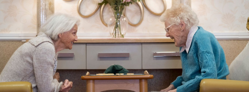 two residents sit across from one another, engaging in a game of scrabble. They are sitting on yellow armchairs, smiling at one another