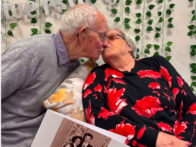 A couple - a man with grey hair, smart clothes, and glasses, leans down to kiss his wife. She is wearing a dark blouse with red flowers. 