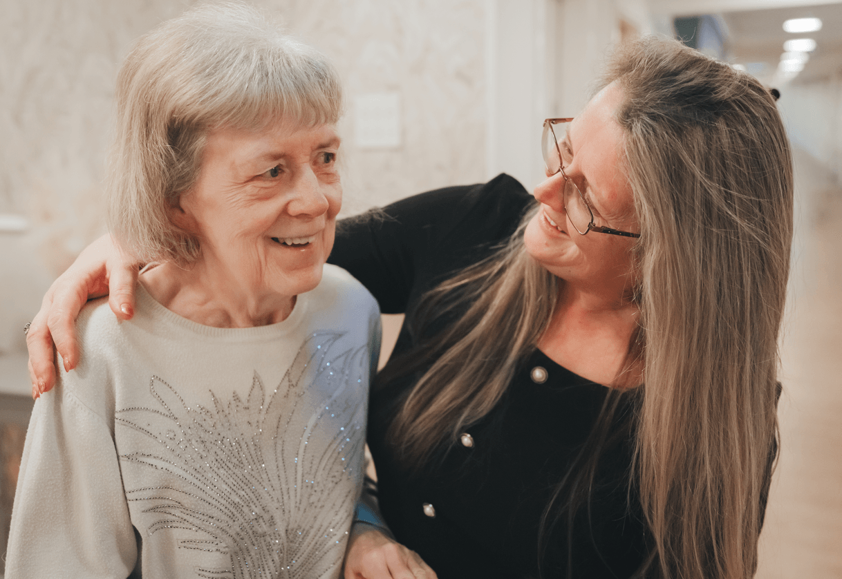 A female staff member with long hair and glasses smiles warmly at a resident. The staff member has a supportive hand on the woman's shoulder. The resident, wearing a light-coloured sweater with a beaded design, looks ahead with a gentle smile.