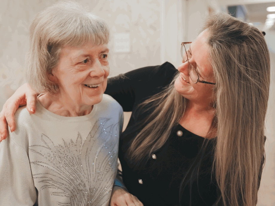 A female staff member with long hair and glasses smiles warmly at a resident. The staff member has a supportive hand on the woman's shoulder. The resident, wearing a light-coloured sweater with a beaded design, looks ahead with a gentle smile.