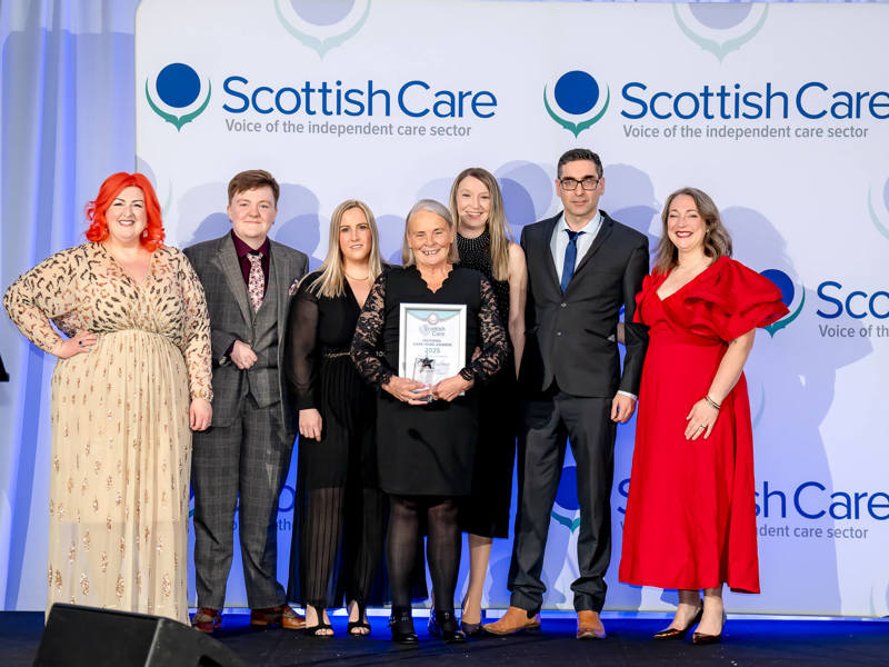 A group of individuals stand on a stage in front of a white backdrop with the "Scottish Care Voice of the independent care sector" logo repeated across it. The woman in the center, dressed in a black dress, holds a framed award certificate.