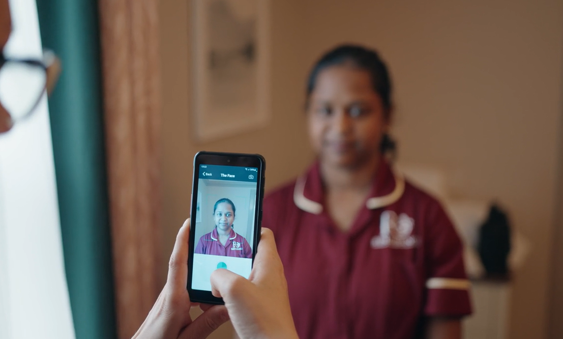 A staff member holds up a nourish device ready to take a photo scan of another staff members face demonstrating the painchek assessment