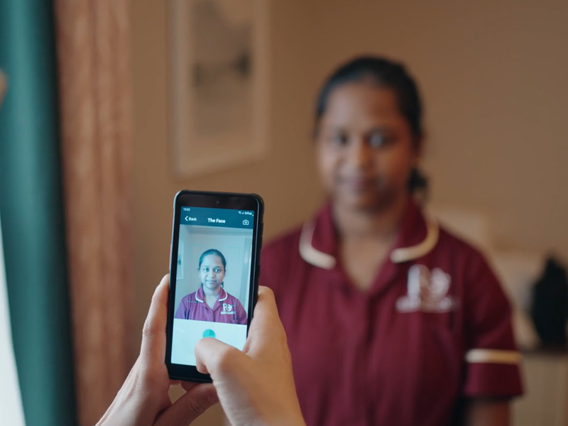 A staff member holds up a nourish device ready to take a photo scan of another staff members face demonstrating the painchek assessment