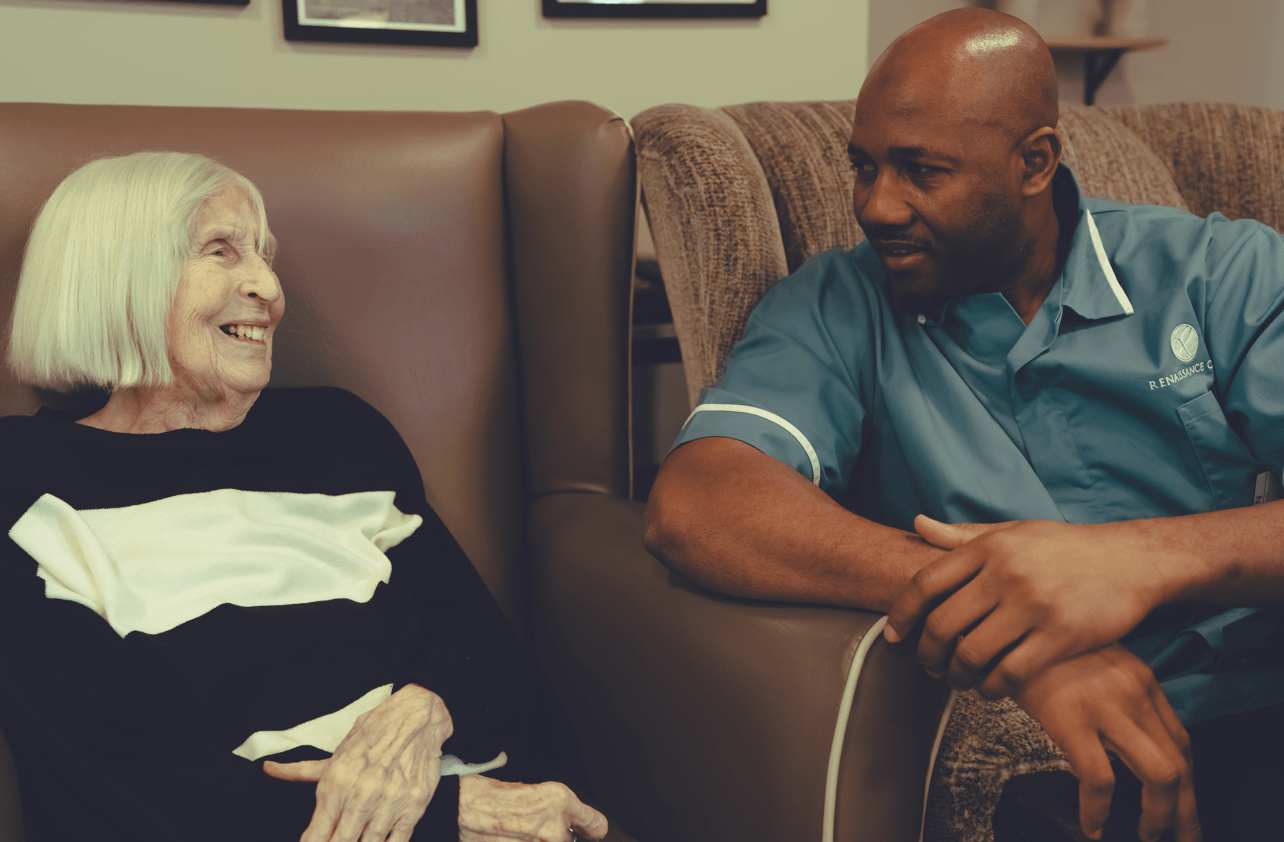 A heartwarming shot of an elderly woman with white hair sitting in a brown leather chair, laughing while talking to a male caregiver. The caregiver is wearing a teal uniform and listening intently.